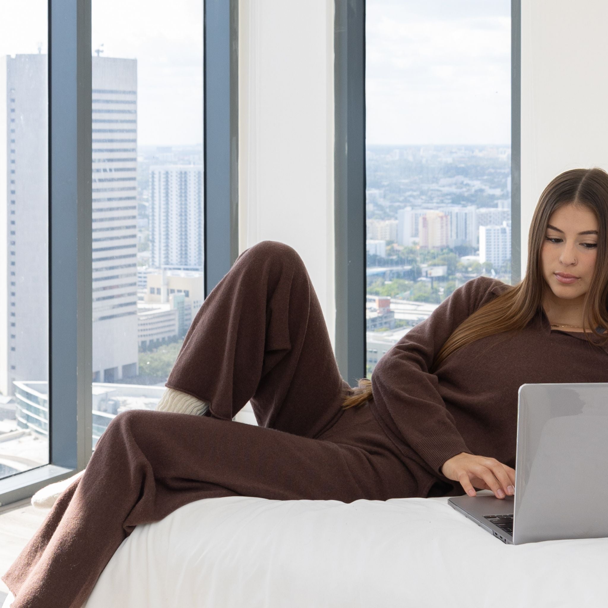 woman laying on a bed in comfortable Smarter at Home set typing on her laptop with the view of a city in the background